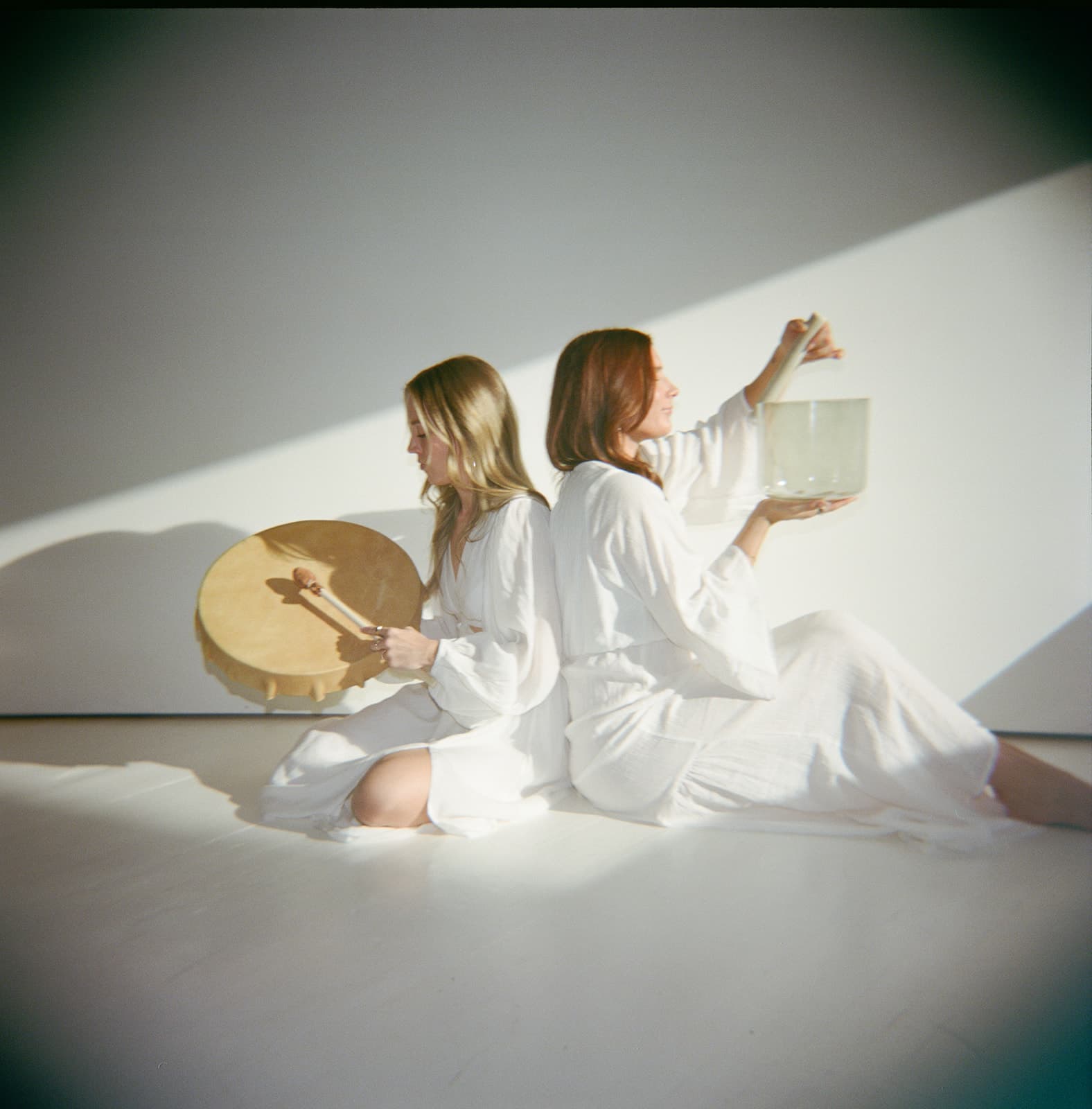 Two facilitators seated back to back with a frame drum and crystal bowl in a bright studio
