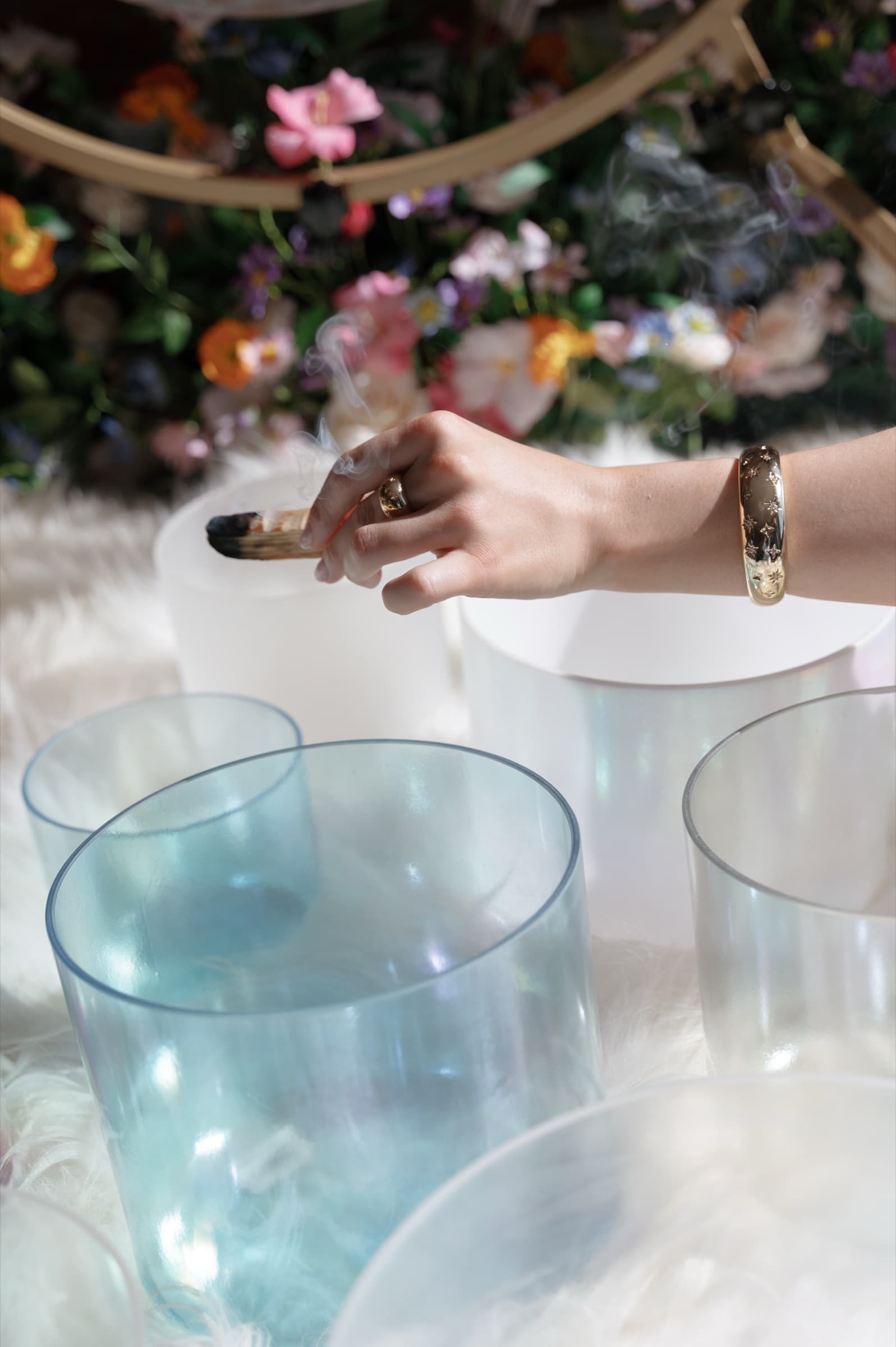 Laura's hand striking a clear crystal singing bowl, with frosted and teal bowls and flowers around it