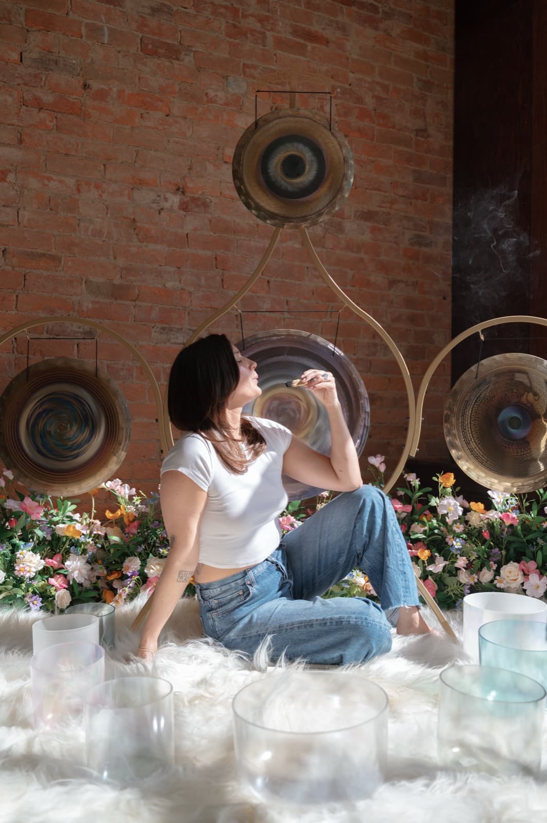 Laura sitting among crystal singing bowls and gongs, eyes closed, in a white tee and jeans