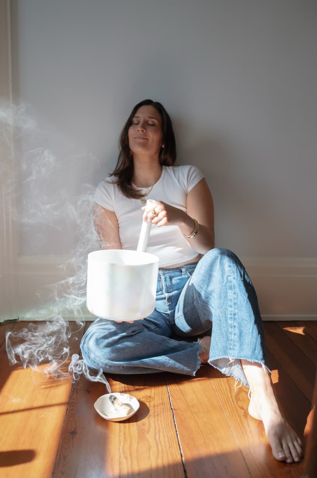 Laura sitting against a white wall in sunlight, playing a crystal singing bowl as smoke curls beside her