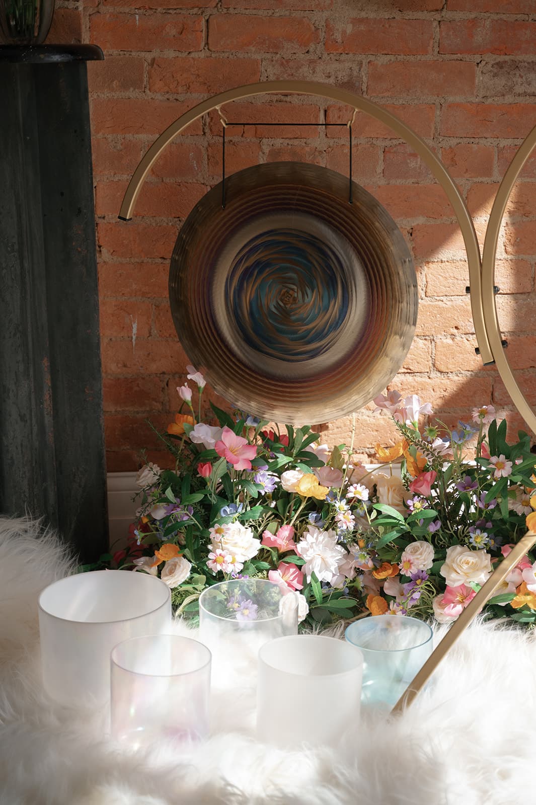 A large gong on a gold stand against exposed brick, banked with flowers above a row of crystal singing bowls on sheepskin
