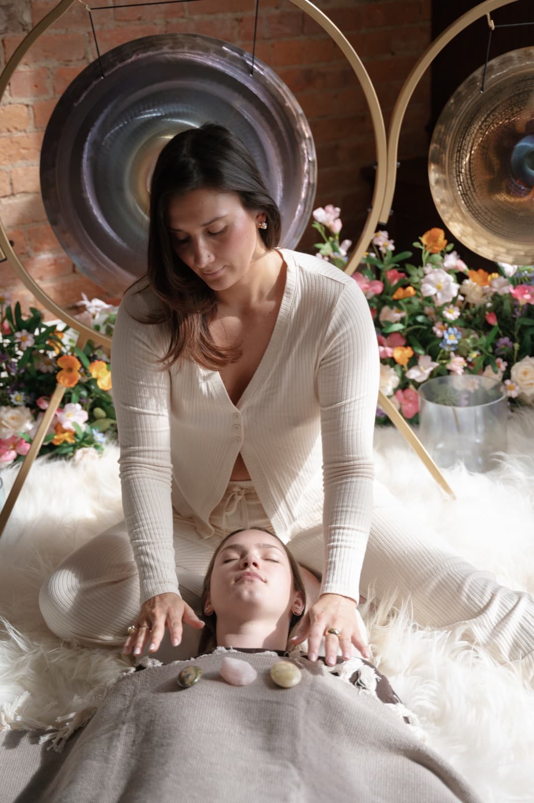 Laura kneeling behind a client with her hands on the client's shoulders, gongs and flowers behind them