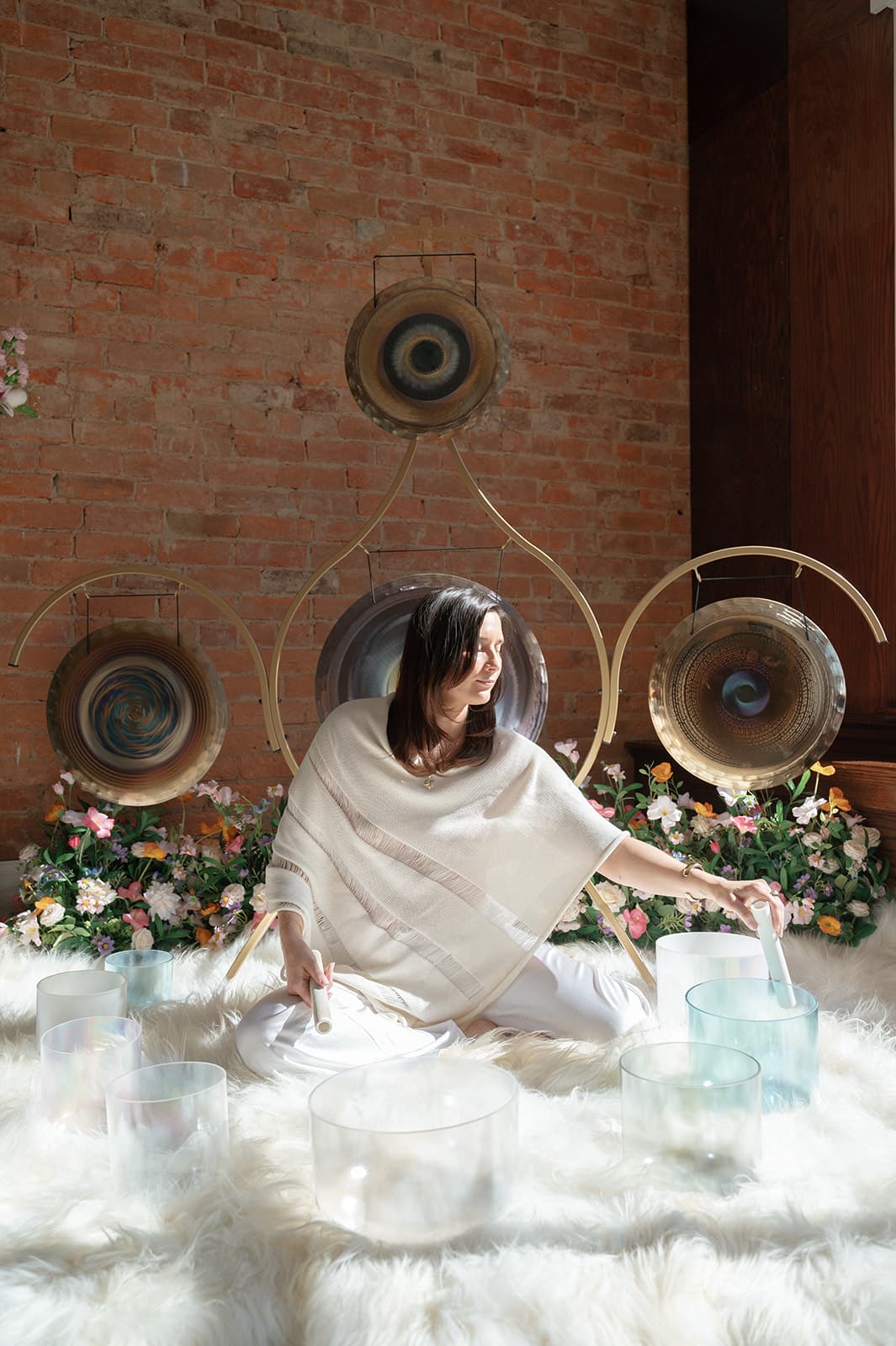 Laura Baker seated on a sheepskin rug, reaching for her crystal singing bowls with gongs and flowers behind her