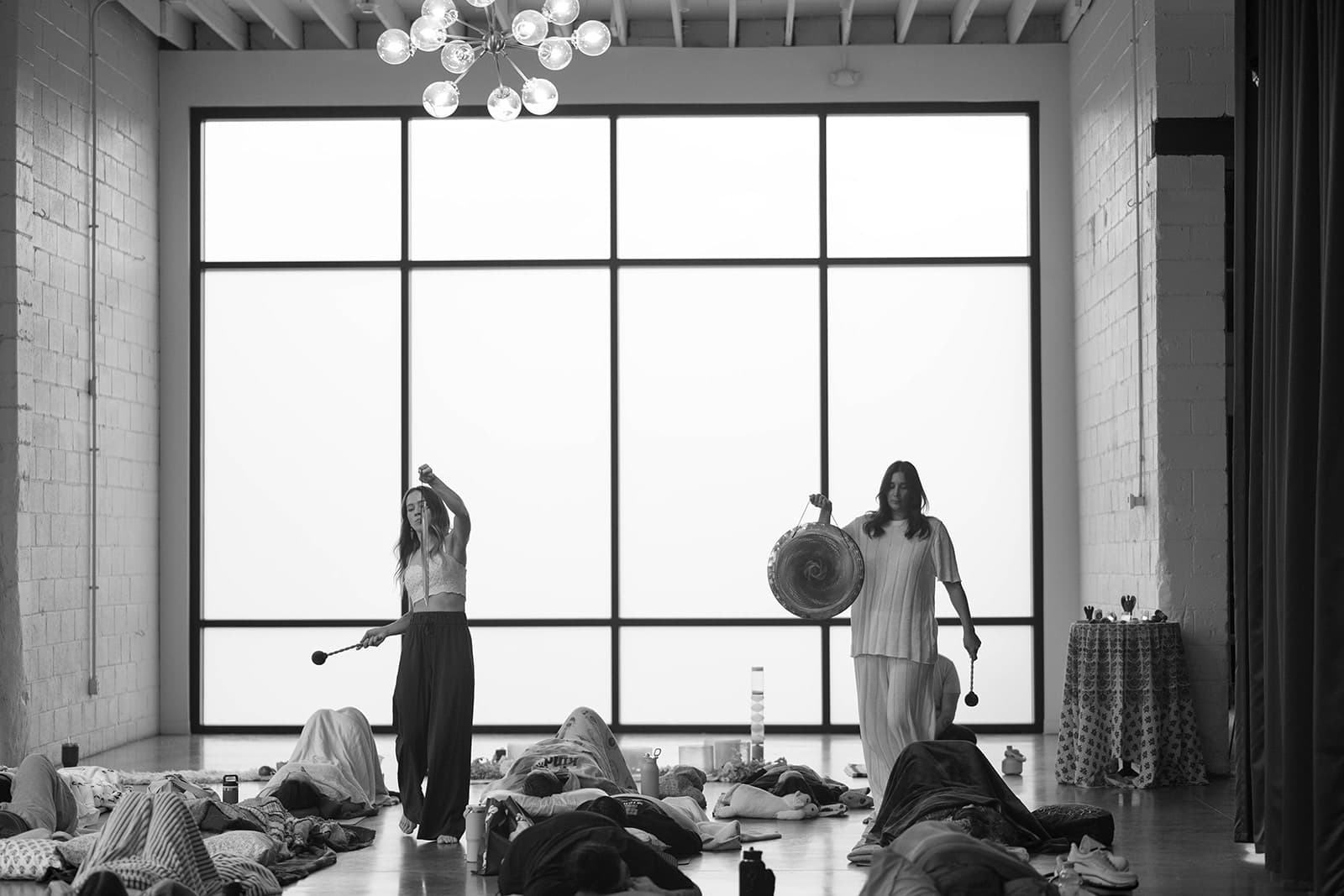 Black and white photo of Laura and Lindsay playing gongs for a room of people resting on mats beneath tall studio windows