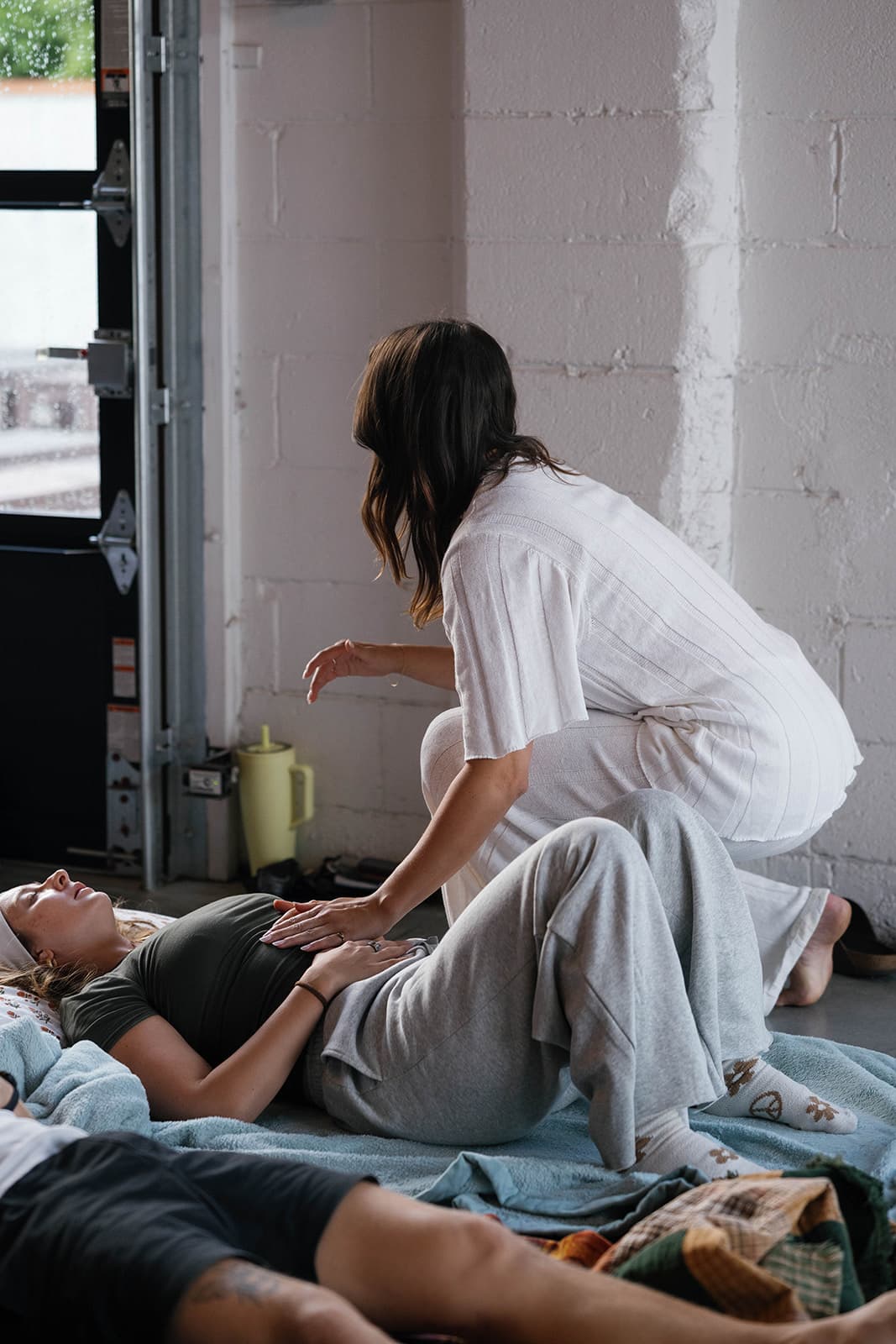 Laura kneeling beside a participant resting on a mat, offering gentle support during a group session