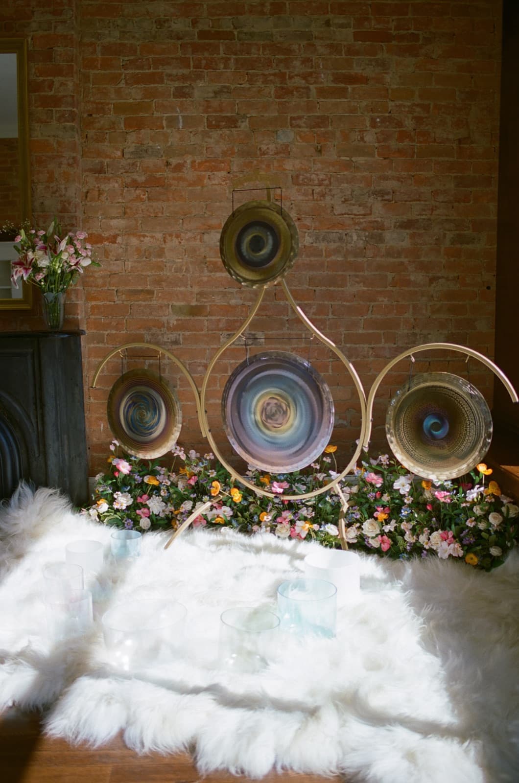 Laura's gongs on a gold stand against exposed brick, with flowers and a sheepskin rug below