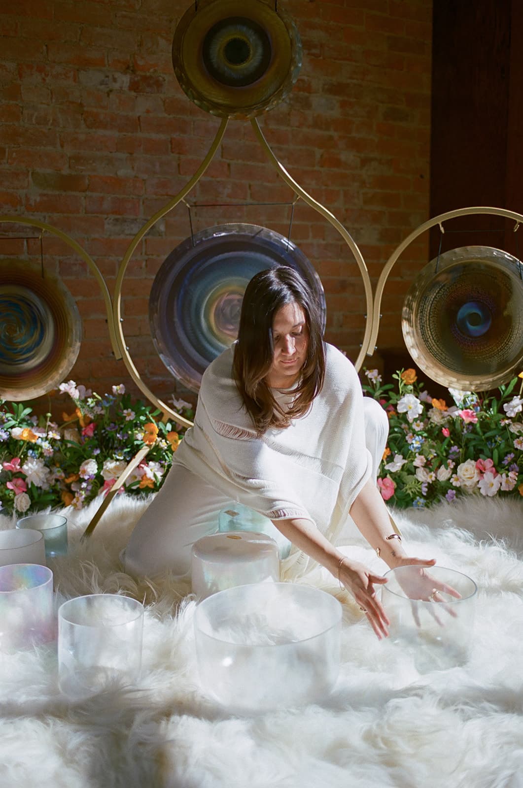 Laura kneeling at her gongs, reaching over crystal singing bowls amid flowers